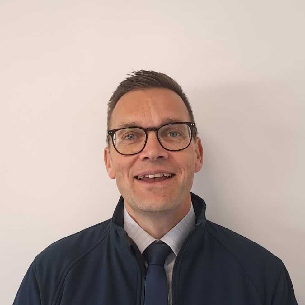 A man wearing glasses and a tie in front of a white wall.